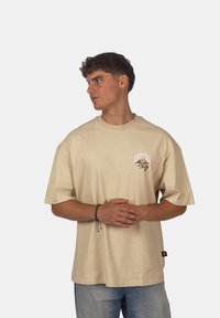 Béžové oversized bavlněné tričko s potiskem loga v hnědé barvě na hrudi, krátkými rukávy a rovně střiženým lemem.