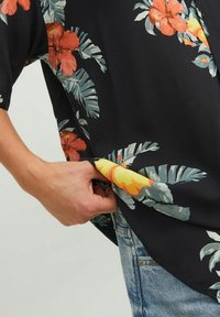 Maglietta nera a maniche corte con un grande motivo floreale in rosso, giallo e verde; tessuto morbido; vestibilità rilassata; mano che tiene l'orlo vicino ai jeans blu.
