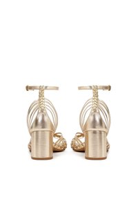 Metāliski zelta krāsas papēža sandales ar bloku papēdi, pītām potīšu siksnām un vairākiem plāniem līkiem siksniņām pāri papēdim, skatītas no aizmugures.