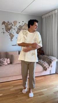 Mann lächelt und verschränkt die Arme, steht barfuß auf einem Holzboden im Wohnzimmer mit beigem Sofa und Wanddekoration in Form einer Weltkarte.