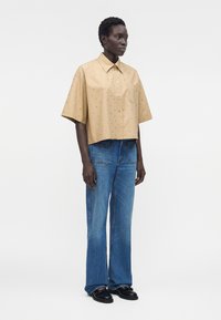 Mujer de pie con una camisa beige de manga corta y suelta con pequeños estampados, vaqueros azules de pierna ancha y zapatos negros, mirando hacia la derecha.