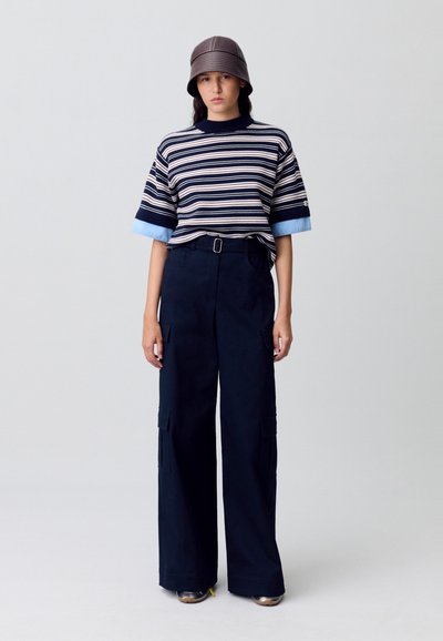 Top de manga corta a rayas en colores azul marino y azul claro; pantalones cargo de pierna ancha en azul marino con bolsillos; sombrero estilo cubo marrón; zapatos plateados.