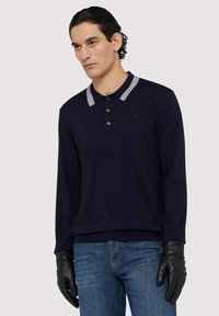 Polo a maniche lunghe di colore blu navy con colletto grigio, tre bottoni e un logo discreto. Abbinato a jeans blu e guanti in pelle nera.
