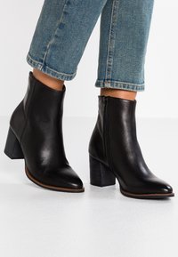 Botines de cuero negro con punta afilada y tacón grueso. Cuentan con un cierre lateral y una textura suave. Combinados con jeans azules recortados.