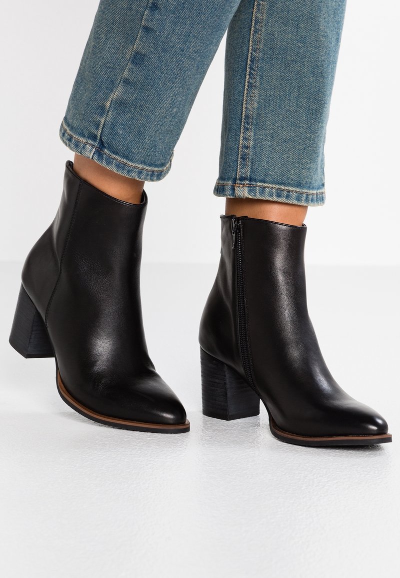 Botines de cuero negro con punta afilada y tacón grueso. Cuentan con un cierre lateral y una textura suave. Combinados con jeans azules recortados.