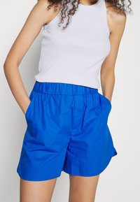 Blue lightweight shorts with an elastic waistband and side pockets, featuring a smooth texture and relaxed fit. Accompanied by a white tank top.