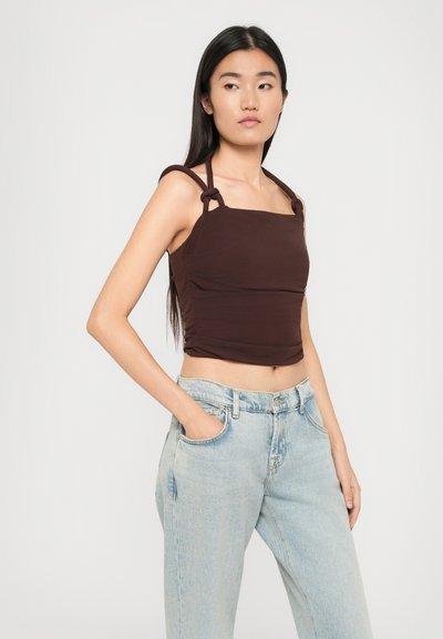 Bruine cropped top met vierkante halslijn, knoopdetail op de bandjes en gerimpelde zijkanten, gecombineerd met lichtblauwe high-waisted jeans.