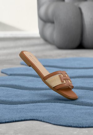 Brązowy klapek na płaskiej podeszwie z plecionym paskiem, leżący na teksturowanym niebieskim dywanie, wnętrze z rozmytym tłem mebli w neutralnych tonacjach.