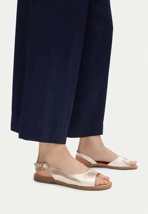 Metallic-goldfarbene Slingback-Sandalen mit offenem Zehenbereich, glatter Textur, verstellbarer Schnalle und flacher brauner Sohle, kombiniert mit marineblauen Hosen mit weitem Bein.