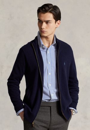 Jeune homme portant un pull zippé bleu marine sur une chemise à rayures bleues et blanches boutonnée et un pantalon gris, regardant vers sa gauche les mains dans les poches.