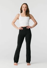 Vrouw staat blootsvoets met handen in haar zij, draagt een wit mouwloos topje en zwarte strakke broek tegen een effen witte achtergrond.