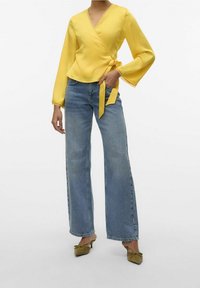 Žlutá blůza s dlouhými rukávy a vlnitými okraji, částečně spárovaná s modrými širokými kalhotami s vysokým pasem a olivově zelenými botami s mašlí.