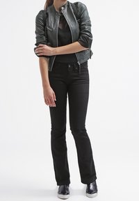 Femme portant une veste en cuir noire, un haut en résille noir, un pantalon évasé noir et des chaussures noires sans lacets, debout les bras croisés.