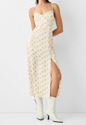 Bloemen midi-jurk in lichtgeel met blauw patroon, aansluitend lijfje, dunne bandjes en een zijsplitsing. Draagt daarbij witte enkellaarsjes met hak.