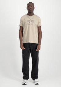 Camiseta de algodón beige con logotipo impreso, combinada con pantalones vaqueros negros. El modelo está en una posición neutral con un fondo claro y zapatillas.