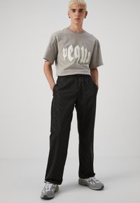 Graues T-Shirt mit großen weißen Schriftzügen, kombiniert mit schwarz gemusterten Hosen. Das Modell trägt graue Sneakers mit weißen Akzenten.