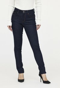 Dunkelblaue Denim-Jeans im Slim-Fit mit einem Fünf-Taschen-Design und kontrastierender Naht. Kombiniert mit einem weißen gerippten Langärmeloberteil.