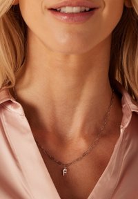 Zilveren ketting met verlengde schakels en een kleine hanger in de vorm van de letter "F," gedragen over een lichtroze satijnen shirt.