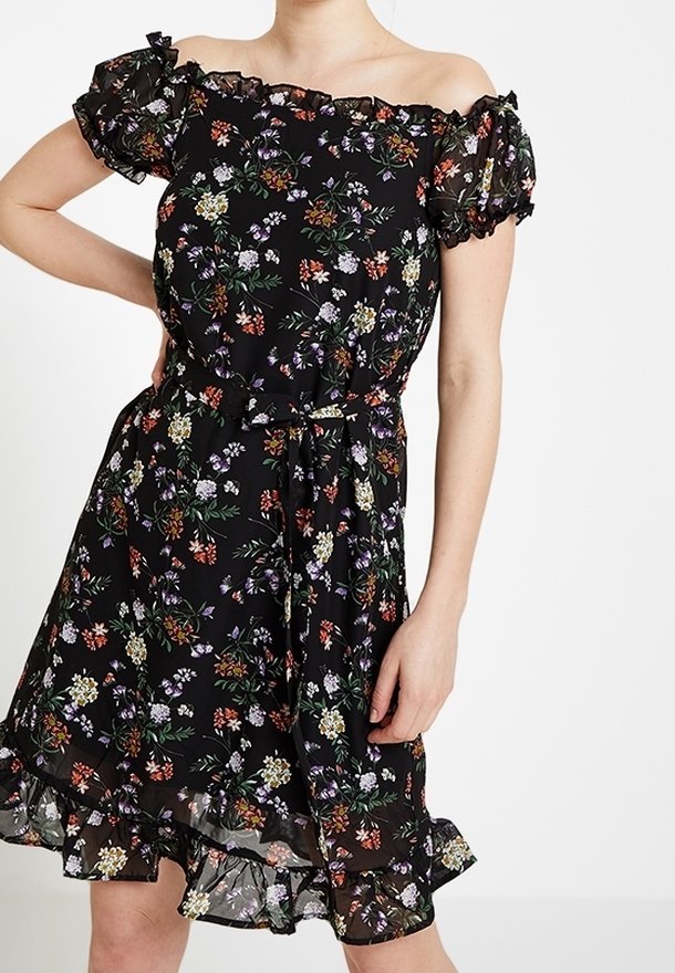 Femme portant une robe noire épaules dénudées avec des manches et un ourlet à volants, décorée d'un petit motif floral multicolore.
