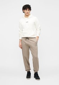 Biała bluza z kapturem, z przodu kieszeń i logo. Beżowe spodnie dresowe z elastycznymi mankietami i sznurkiem. Czarne trampki.