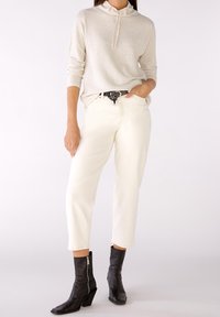 Jasnobeżowy sweter z kapturem, kremowe zwężane dżinsy oraz czarne botki za kostkę na suwak, z czarno-białym paskiem w kropki.