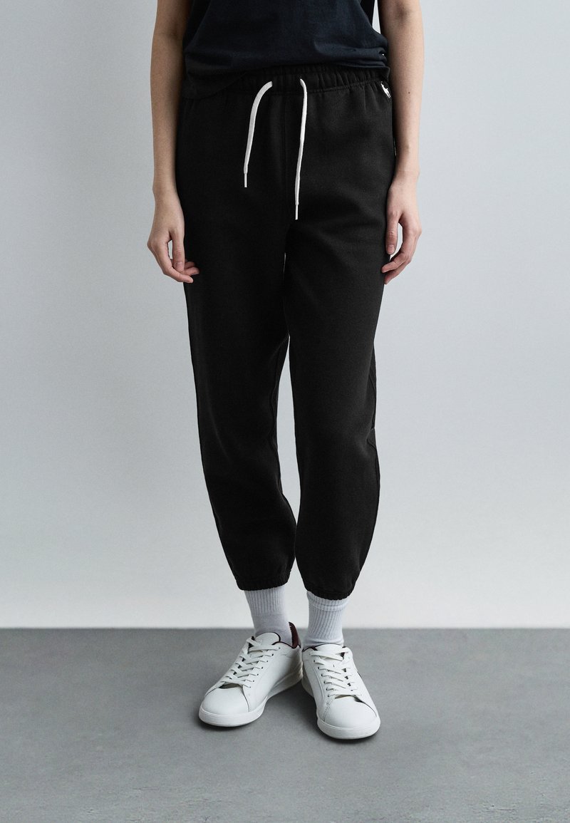 Άτομο που φοράει μαύρο παντελόνι φόρμας με λευκά κορδόνια, λευκές κάλτσες και λευκά αθλητικά παπούτσια, στέκεται σε γκρίζο πάτωμα μπροστά σε λευκό τοίχο.