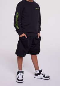Black sweatshirt with neon green branding on the sleeve, paired with black mesh shorts and black-and-white high-top sneakers.