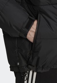 Svart puffjacka med en dragkedjeficka och velcro-stängning vid ärmarna. Har en mjuk textur och draperad fåll. Värmande isolerad design.