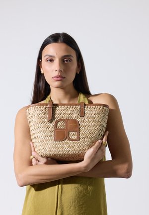 Jeune femme aux longs cheveux foncés portant une robe olive sans manches tient un sac à main tissé beige avec des détails en cuir marron.