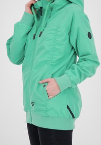 Veste vert menthe avec capuche, fermeture éclair sur le devant, cordons de serrage et poignets élastiques. Elle dispose de poches latérales et d'un logo en forme de cœur près de l'ourlet.