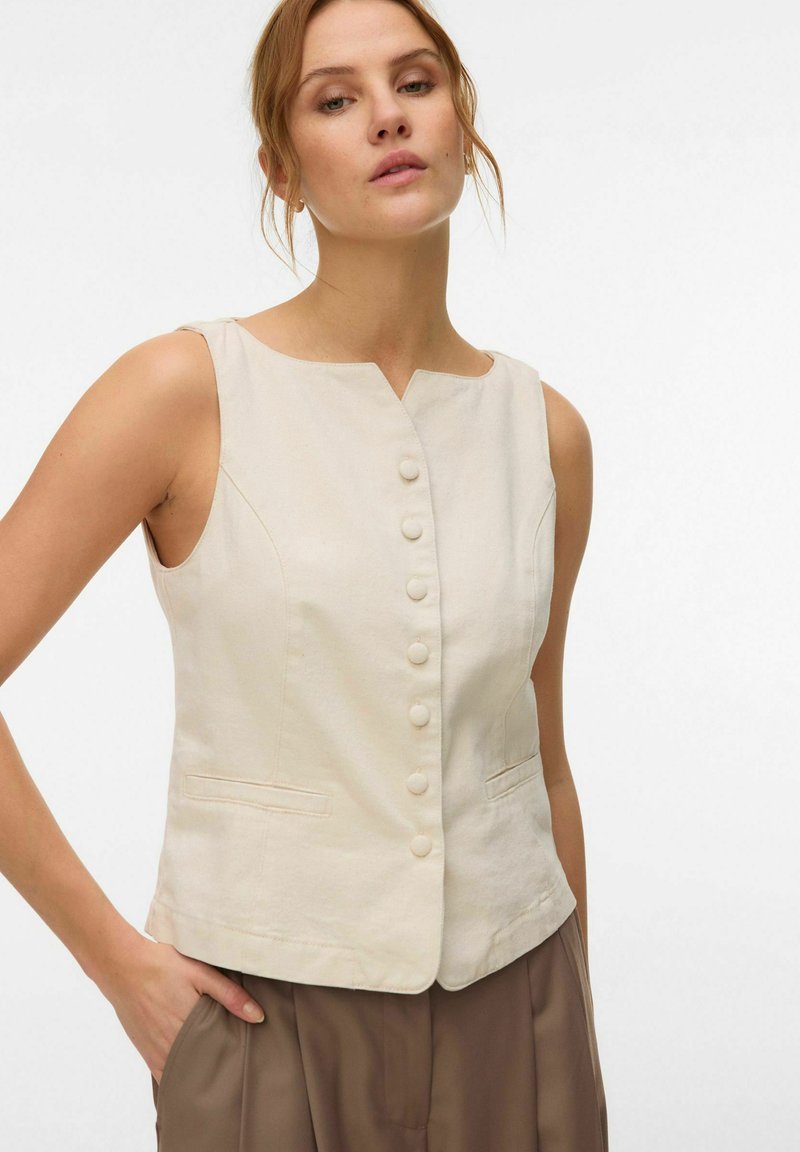 Femme portant un gilet beige sans manches boutonné sur un pantalon marron, debout avec une main dans la poche sur un fond blanc uni.