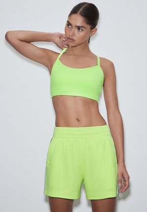 Jeune femme en crop top vert néon et short assorti, posant avec un bras plié derrière la tête sur un fond clair uni.