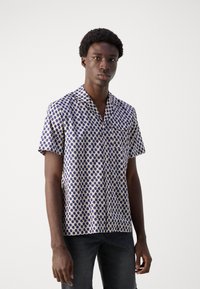 Camisa de manga corta hecha de tejido ligero, con un patrón de lunares azules sobre un fondo crema, con un cuello puntiagudo y un bolsillo en el pecho.