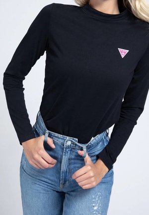 Kvinde iført sort langærmet top med pink Guess-logo og højtaljede blå jeans, hænder justerer linningen.