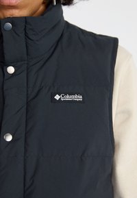 Colete isolante preto com colar alto, botões de pressão e um patch do logótipo da Columbia no lado esquerdo da frente. Tecido suave.