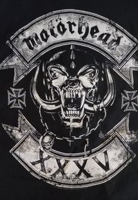 Svart T-skjorte med et slitt bilde av en fryktinngytende hodeskalle med støttenner, flankert av kors og teksten "motorhead" over.