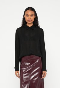 Melna svītraina blūze ar pogām priekšpusē un apkakli, kopā ar bordo krāsas spīdīgu svārku ar priekšējo šķēlumu un paneļu dizainu.