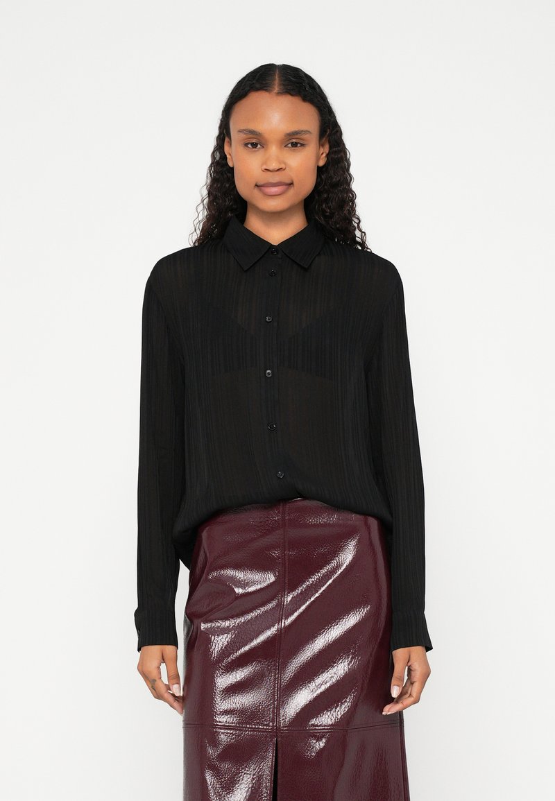 Melna svītraina blūze ar pogām priekšpusē un apkakli, kopā ar bordo krāsas spīdīgu svārku ar priekšējo šķēlumu un paneļu dizainu.