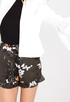 Femme portant un haut blanc à manches longues bouffantes et un short noir à volants fleuris sur un fond uni.