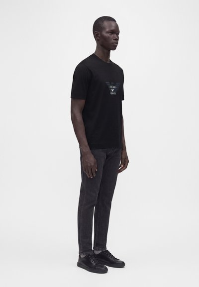 Άνδρας που στέκεται φορώντας μαύρο T-shirt Emporio Armani, σκουρόχρωμο τζιν και μαύρα αθλητικά παπούτσια μπροστά σε απλό λευκό φόντο.