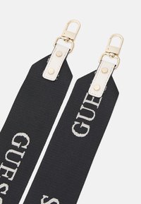 Sangle en tissu noir avec le logo "GUESS" en blanc, équipée de ferrures dorées et d'accents en cuir. Les extrémités comportent des attaches à fermoir et des boutons-pression.