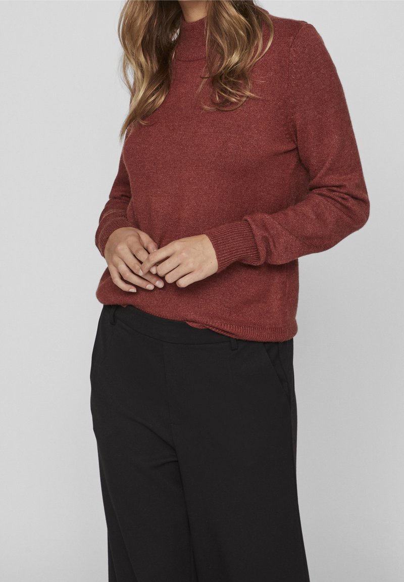 Femme portant un pull en maille couleur rouille et un pantalon noir taille haute, les mains jointes légèrement à la taille, cheveux ondulés clairs visibles.