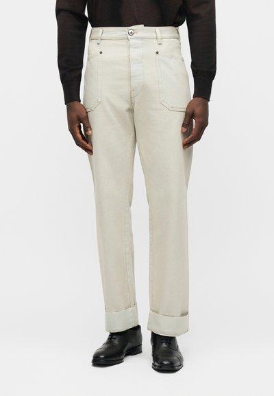 Άνδρας με κρεμ ευθείς παντελόνια, μεγάλα μπροστινά τσέπες, γυρισμένα τελειώματα, μαύρα παπούτσια και μακρυμάνικη μαύρη μπλούζα.