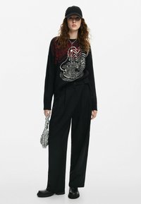 Maglione nero oversize in maglia con design astratto rosso e bianco, abbinato a pantaloni neri a vita alta, borsa argentata con texture e stivali neri.