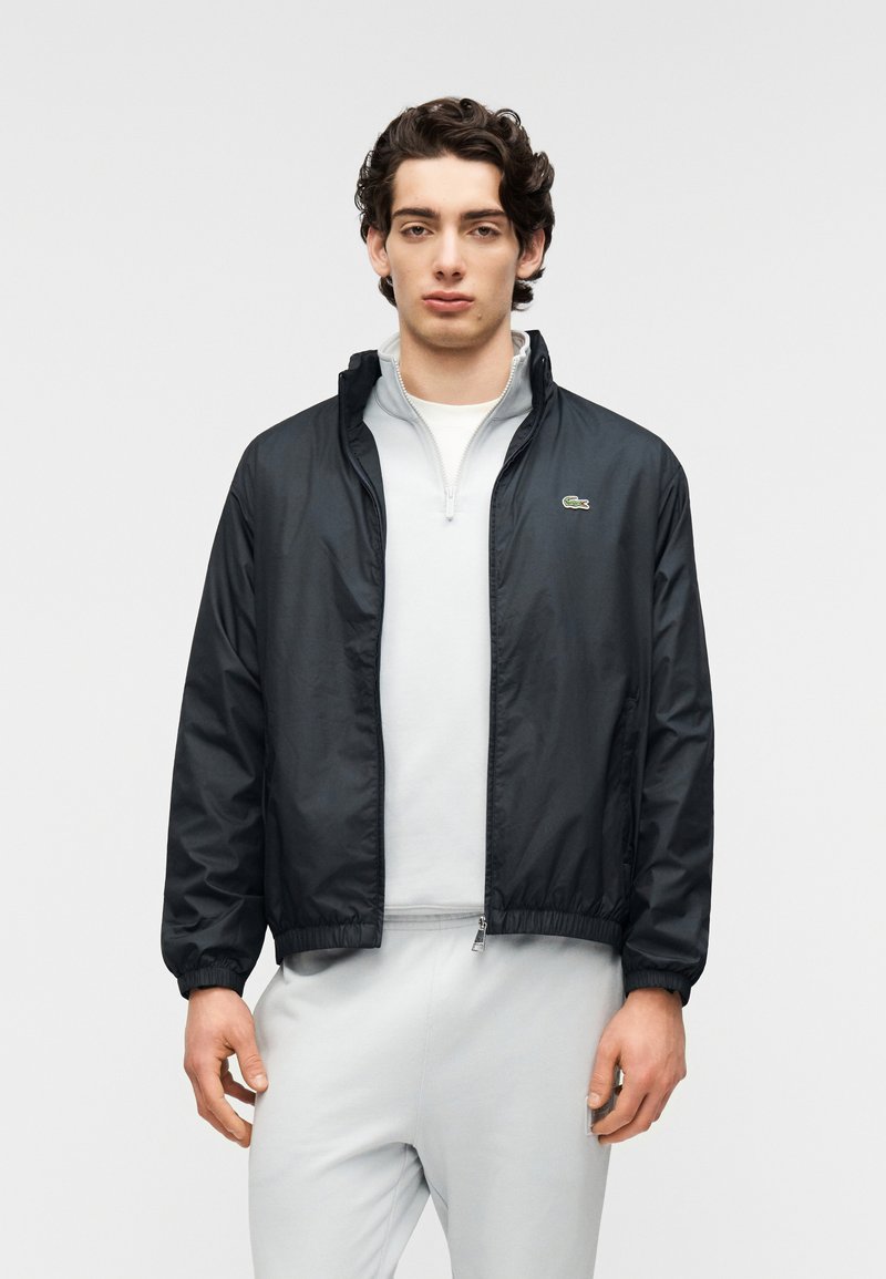 Μαύρο ελαφρύ μπουφάν με ελαστικές μανσέτες, φερμουάρ μπροστά και λογότυπο Lacoste. Φορεμένο πάνω από γκρι ανοιχτόχρωμο τοπ με φερμουάρ. Λείο, ανθεκτικό στο νερό υλικό.