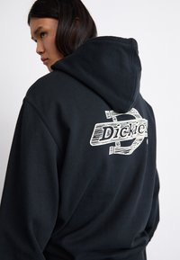 Sort hættebluse med et stort hvidt logo på ryggen med teksten "Dickies." Stoffet ser blødt ud, med en struktureret, hyggelig fremtoning og snørebånd i hætten.