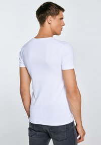 Fiatal férfi oldalra nézve, szűk fehér rövid ujjú inget és sötétszürke farmert viselve, egyszerű háttér előtt.