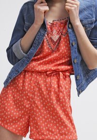 Czerwony kombinezon w wzory z troczkami w talii, założony pod niebieską kurtką dżinsową z złotymi guzikami i podwiniętymi rękawami.