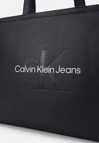 Crna torba od umjetne kože s reljefnim logotipima 'CK' i 'Calvin Klein Jeans' u bijeloj boji. Pravokutni oblik s dva gornja ručka.