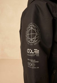 Schwarze Jacke mit strukturiertem Stoff und gedruckten Grafiken auf dem Ärmel, darunter ein Globusdesign und alphanumerischer Text.